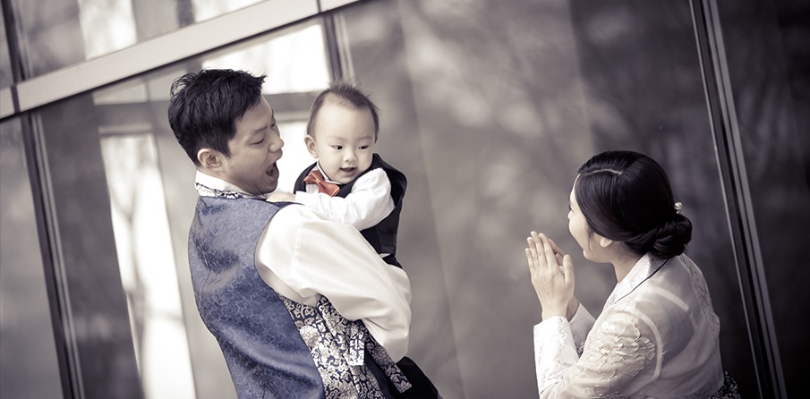 야외 공간 — 아빠 품의 수트 차림 아기, 엄마가 손뼉 치며 반응 유도