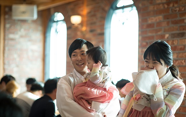 실내 홀 벽돌벽과 아치창 배경 — 아빠가 하랑이 안고 엄마와 함께