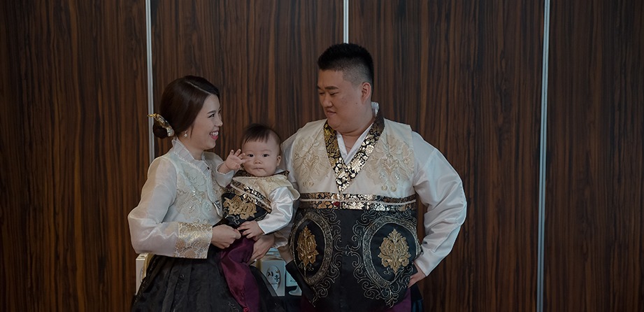 실내 나무벽 앞 가족 3인 — 아기가 손을 들어 눈 가리는 순간